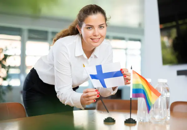 身穿商务服装的年轻女性将芬兰国旗和男女同性恋 双性恋和变性者的旗帜摆上谈判桌 — 图库照片