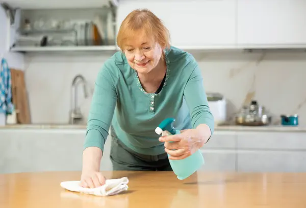 Hardworking mature woman cleaning the kitchen at home with cleaning products wipes the table with a damp cloth