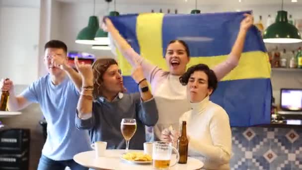 幸せな友人のグループ スポーツサポーターは パブでテレビで試合を見ながらスウェーデンの旗を持つお気に入りのチームを応援 — ストック動画