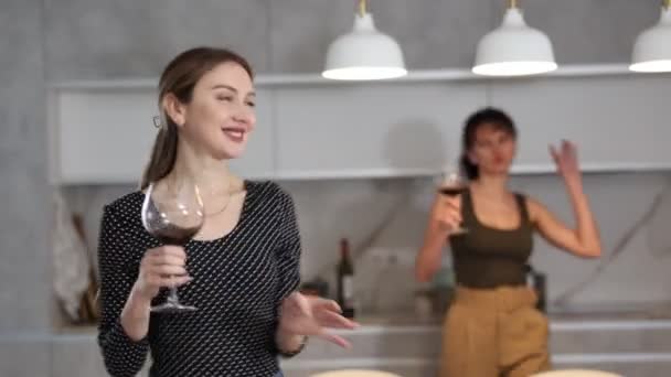 女性はワインを飲んでいて キッチンで音楽を踊っています 背景では 彼女の手のワインのグラスと友人は動きを繰り返す — ストック動画