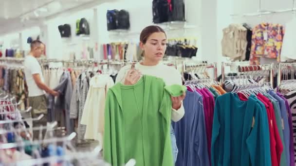 Zadowolona Młoda Dziewczyna Wybiera Jednokolorowy Sweter Sklepie Odzieżowym Dużym Asortymentem — Wideo stockowe