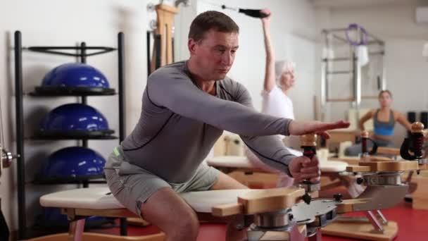 ジムでは 男性は体重を減らすためにピラティス演習を行い ヘロンプーリータワーを使用して腕 体の筋肉をもたらします 全身ポンプ用総合機器 — ストック動画
