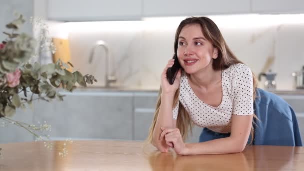 ホワイトインテリアのキッチンでテーブルの上に置かれている電話で話すジョヨウス中年の女性 — ストック動画