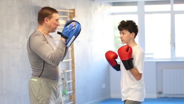 男孩戴着拳击手套 练习拳击 冲着垫子打拳击 十几岁的男孩和她的教练父亲在体育馆里 高质量的4K镜头 — 图库视频影像
