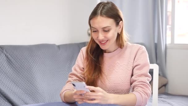 坐在公寓桌子旁边笑着的年轻女人在用手机发短信 是的高质量的4K镜头 — 图库视频影像