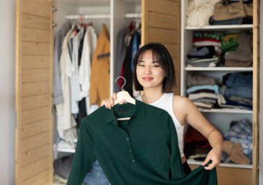 Glad young Asian woman looking at dresses held in hand for choice in dressing room clipart