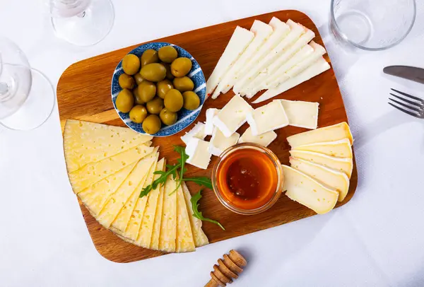 在白色的桌子上有切菜板 上面放着各种奶酪 腌制橄榄 轻质酒精饮料的精美小吃 奶酪切片用牛油果叶装饰 — 图库照片