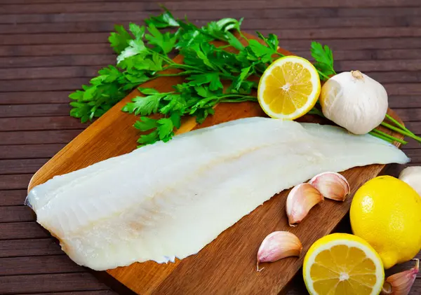 Image of fillet of raw cod fillet before cooking on wooden background with garlic and greens