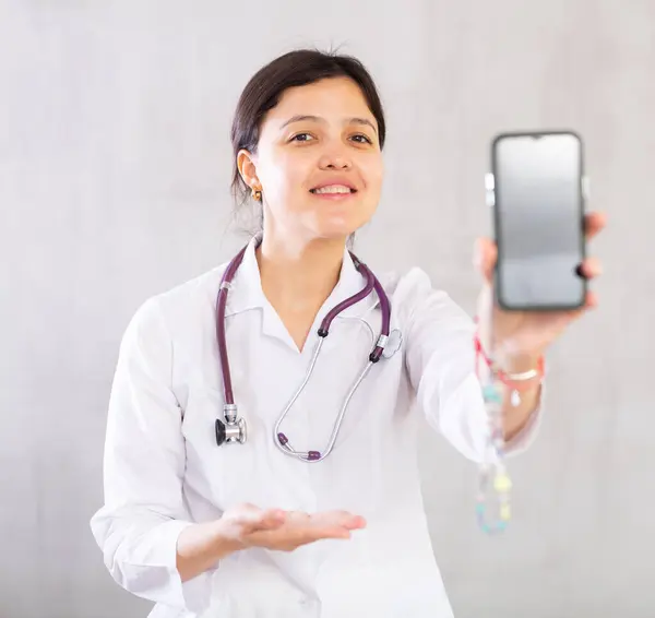 身穿白衣 面带微笑的年轻女医生推荐移动医疗应用 女人伸出手与智能手机 展示小玩意屏幕 — 图库照片