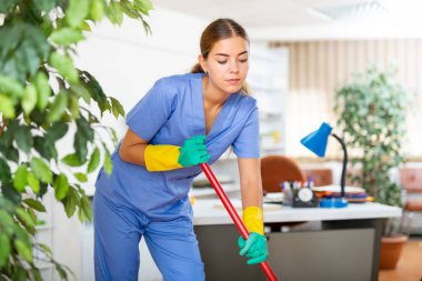 Portrait of professional female worker of office cleaning service wearing uniform and rubber gloves wiping floors with mop clipart