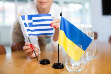 Female hands arranging flags of Greece and Ukraine on table before international summit meeting, symbolizing importance of relations between countries. Selective focus clipart