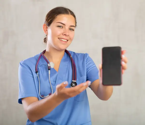 Smiling female nurse in blue uniform recommending mobile medical app, stretching out hand with smartphone. Focus on device screen
