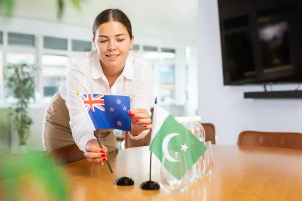 ポジティブな若い女性は 会議室のパキスタンの旗と水のボトルの隣のテーブルにオーストラリアの小さな旗を置く — ストック写真