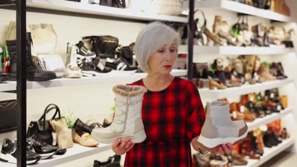 シニアの女性バイヤーは新しいコレクションの履物を見て 冬のための新しいブーツをピックアップします マルチブランドの店では 顧客は手で靴の展示サンプルを保持し 購入について考えます — ストック動画
