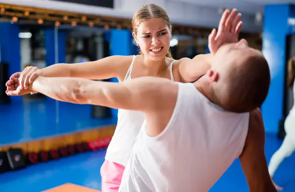 Young woman practicing basic self-defense techniques while training in gym with male partner, performing palm heel strike in chin