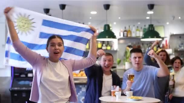 パブで一緒に試合を見ながら お気に入りのチームとウルグアイの旗を振っている若い大人のスポーツファンを叫ぶ — ストック動画