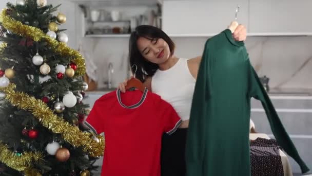 年轻的亚洲女人看到手中拿着可供选择的衣服 站在自家圣诞树旁 感到很高兴 但并不满意 — 图库视频影像