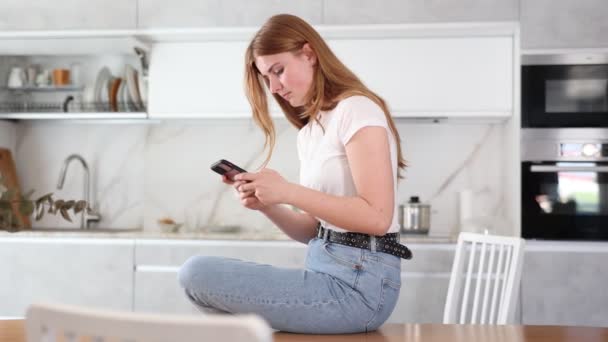 白いTシャツとジーンズの悲しい少女はキッチンテーブルに座り ソーシャルネットワーク上で彼女のボーイフレンドと通信する — ストック動画