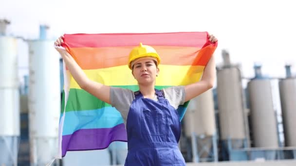 ヘルメットのポジティブな若い女性労働者は 晴れた夏の日に精製工場で大きなタンクの前に立っている間 虹Lgbtの旗を振ります — ストック動画
