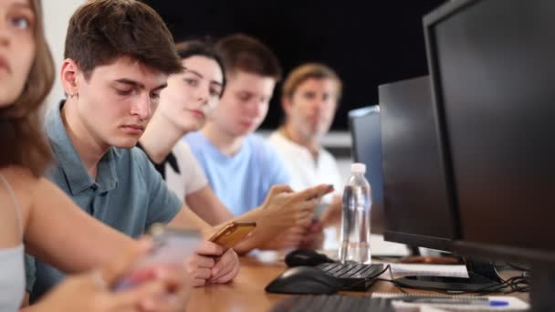 授業中のコンピュータスクールでは 若い男の子の学生が携帯電話で座り 教師に敬意を表してリストします — ストック動画