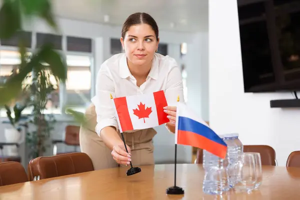 stock image Young woman in business clothes puts flags of Canada and Russia on negotiating table in office