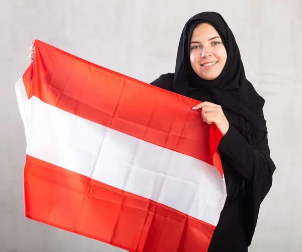 陽気な若いイスラム教徒の女性は 黒の伝統的なヒジャブを着て オーストリアの旗を保持しています ストックフォト