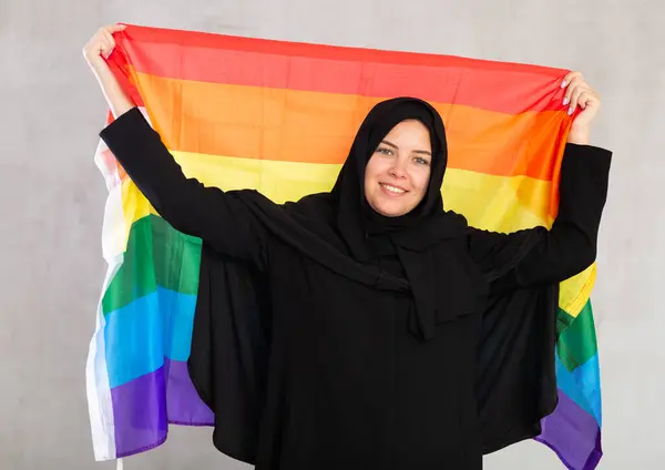 Lgbtプライドの旗を保持する黒の伝統的なヒジャブを身に着けている陽気な若いイスラム教徒の女性 ロイヤリティフリーのストック画像