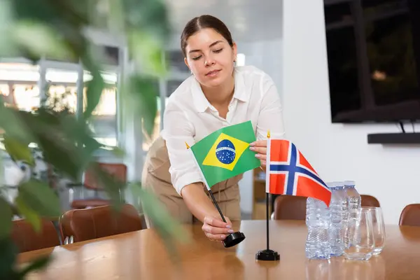 ポジティブな若い女性は 国際交渉のための会議室でノルウェーの旗とテーブルにブラジルの小さな旗を置く — ストック写真