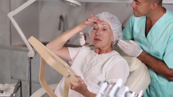 男性のプラスチック外科医は 診療所で鏡を見ているシニア女性顧客に顔の輪郭を描いた後に専門的なアドバイスを与えます — ストック動画
