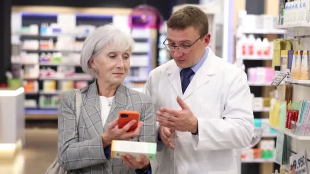 高齢の女性は 彼女が彼女のスマートフォン画面で電子処方箋を読むのを助けるために薬剤師に頼みます 高品質の4K映像 — ストック動画