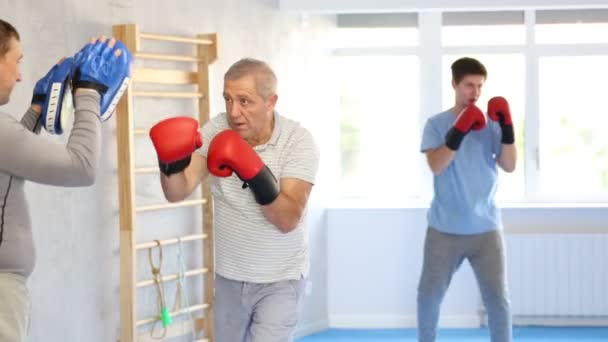 在训练室的联合训练中 对拳击手套感兴趣的年长男子在成年儿子手中的手套上打拳 高质量的4K镜头 — 图库视频影像