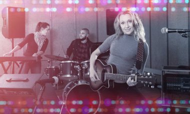 Portrait of excited young blonde girl rock singer with guitar during rehearsal with male drummer and female keyboardist in studio clipart