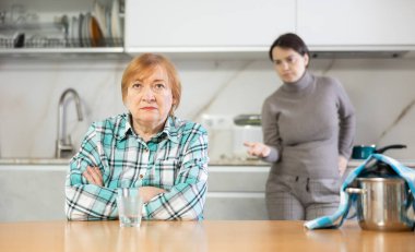 Young girl makes claims her mother emotionally.Sad mature woman with soup in pan on table listens to reproaches of daughter sitting on kitchen at home. Misunderstanding relationship clipart