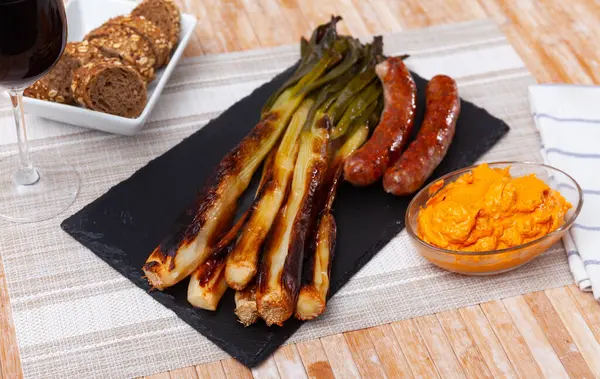 stock image Traditional Catalan feast with roasted calcots served on slate board with rich romesco sauce, grilled butifarra sausage, multigrain bread and glass of wine in casual dining setup..