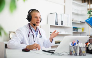 Professional therapist in headphones advising remote patient online via video call on laptop while sitting in medical office clipart