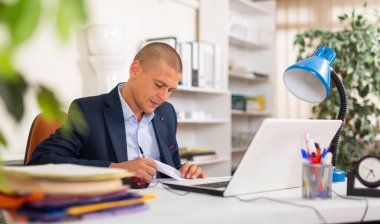 Positive businessman working with laptop and papers at office desk clipart