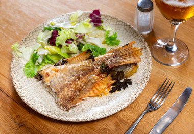 Piece of baked perch with salad of fresh vegetables is served on plate. Baked fish dish is garnished with pieces of fresh vegetables, complemented with glass of beer clipart