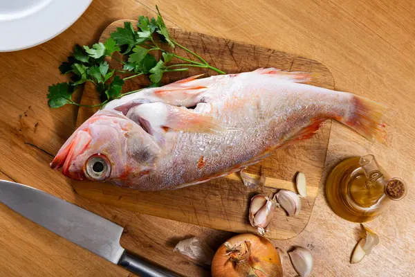 stock image Fresh whole raw alfonsino fish prepared for cooking on wooden kitchen table accompanied by green parsley, garlic cloves, onion, and olive oil