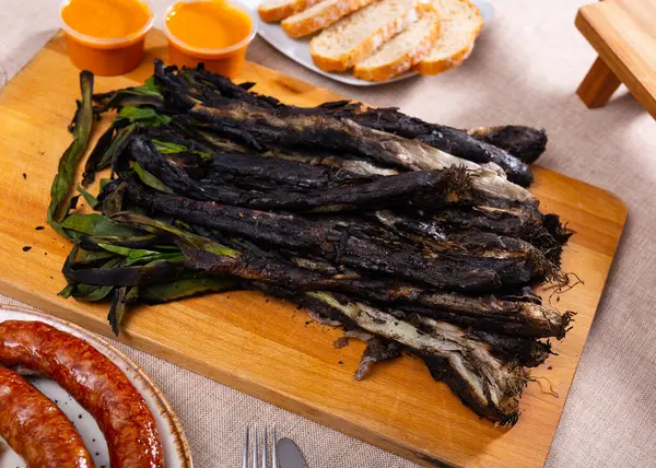 stock image Traditional dishes of Catalonia, calcot with romesco sauce served on wooden board, bread and grilled butifarra sausage on table