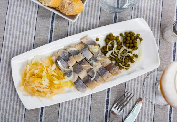 stock image Mouthwatering Scandinavian appetizer of lightly salted herring filets served on plate with onions and vegetable garnish of pickled cabbage and peas