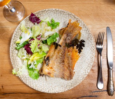 Piece of baked perch with side dish of fresh vegetables is served on plate. Fish is guaranteed with salad of lettuce and parsley, complemented by glass of beer. clipart