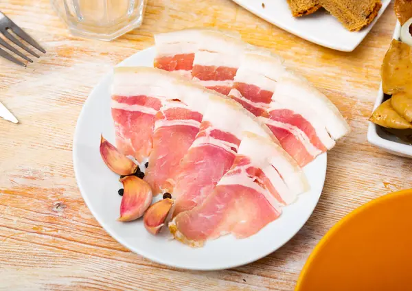 stock image Delicious juicy slices of fresh sliced bacon on a plate with garlic cloves and pepper peas