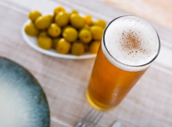 stock image Glass of refreshing lager beer with foamy head, paired with plate of piquant marinated green olives, offering harmonious blend of flavors for appetizer