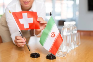 Little flag of Iran on table with bottles of water and flag of Switzerland put next to it by positive young woman  clipart