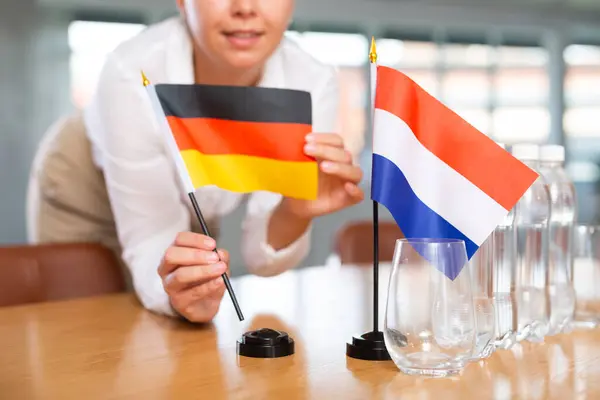 stock image Unrecognizable woman preparing room for international negotiations and communication discussions of leaders. Lady sets miniatures flags of Netherlands and Germany on table. Unfocused shot