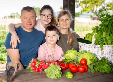 Portrait of smiling parents with two teenage children sitting outdoors at table full of freshly harvested vegetables in backyard garden. clipart