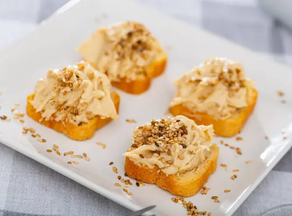 stock image On crispy toast, toast spread with hummus is lean vegetarian dish of Oriental cuisine, made from mashed chickpeas, spices, garlic. Sandwiches decorated with sprinkling of white and black sesame seeds