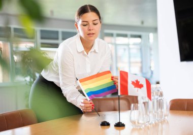 Assistant girl prepares office for international negotiations and meetings of leaders. Lady sets miniatures flags of Canada and LGBT on table. Close-up clipart