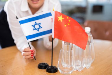 Unrecognizable woman preparing room for international negotiations and communication discussions of leaders. Lady sets miniatures flags of China and Israel on table. Unfocused shot clipart