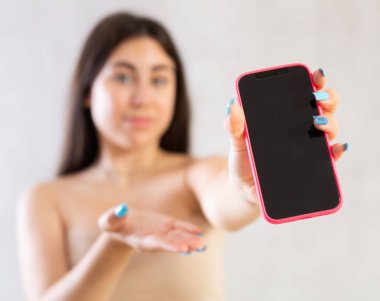 Mobile phone held in hand against blurred background of young woman in front of gray wall clipart